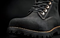 Close-up of rugged black suede work boot with thick rubber sole, metal eyelets and contrast stitching on dark background, showcasing durable construction details.