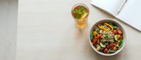 Fresh grain bowl with colorful vegetables, cherry tomatoes, greens, and seeds beside iced herbal tea and open notebook on light wood desk.