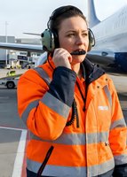 Female airport ground crew worker in orange safety vest using headset radio communication at busy commercial airport terminal with aircraft background.