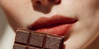 Close-up of person with fair skin biting dark chocolate bar, showing red-tinted lips against pale complexion in soft natural lighting.
