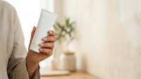 Person with medium skin tone holding blank white cosmetic tube or skincare product container for branding and packaging mockup purposes.