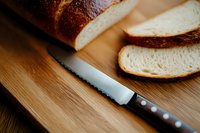 Fresh sliced artisanal bread loaf on wooden cutting board with serrated knife, showing soft white crumb and golden brown crust in warm lighting.