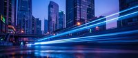 Urban night scene with light trails from moving traffic creating dynamic blue streaks through modern city downtown district with illuminated skyscrapers and office buildings.