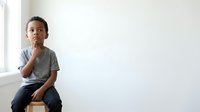 Young boy with dark hair and medium skin tone, wearing casual gray t-shirt and jeans, sitting thoughtfully with hand to chin against white wall background, expressing contemplation and decision-making for educational and lifestyle concepts.