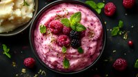 Fresh raspberry smoothie bowl topped with mint leaves and berries, garnished with seeds on dark background, creamy pink texture for healthy breakfast.
