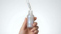 Hand holding frosted glass dropper bottle with silver cap and pipette for skincare serum or essential oil cosmetic product on white background.