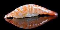 Fresh shrimp nigiri sushi piece on black reflective surface with mirror reflection, showcasing traditional Japanese cuisine preparation and presentation style.