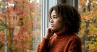 Young African American woman in orange sweater looking thoughtfully out window during autumn season with colorful fall foliage background.