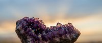 Amethyst crystal cluster glowing against a dramatic sunset sky, showcasing deep purple gemstone formations with natural light rays in the background.