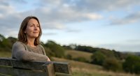 Mature Caucasian woman in beige sweater sitting peacefully on wooden bench outdoors during golden hour, contemplating scenic countryside landscape.