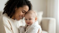 Happy African American mother holding smiling baby in cozy home setting. Tender maternal bonding moment with infant child showing love and care.