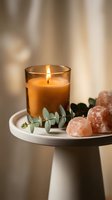 Burning candle with pink salt crystals and eucalyptus on white tray creating peaceful spa atmosphere for relaxation and wellness rituals.