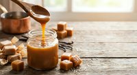 Homemade salted caramel sauce dripping from wooden spoon into glass jar on rustic kitchen counter with caramel candies and copper pot in warm sunlight.