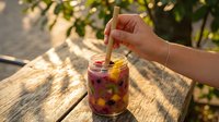 A hand holding a wooden straw in a glass jar filled with berry-infused beverage on a wooden table outdoors, surrounded by natural sunlight and foliage.