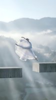 Young woman in flowing white dress leaping between concrete platforms with misty mountain landscape background creating dreamy ethereal atmosphere.