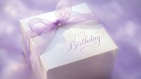 Elegant white gift box with purple ribbon bow and Happy Birthday inscription on soft lavender background for greeting cards.
