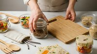 Caucasian female hands pouring dried grains into glass jar on wooden cutting board surrounded by fresh vegetables and ingredients for meal preparation and healthy cooking.