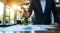 Businessman analyzing financial charts and graphs on desk with sunlight through window, pointing at data during morning strategy meeting.