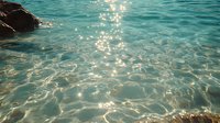 Sparkling turquoise ocean water with sunlight reflections creating shimmering patterns on crystal clear surface, dark rocks visible through transparent shallow sea.