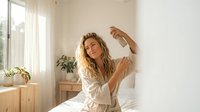 Young blonde woman stretching arms while waking up in bright bedroom with natural sunlight streaming through window creating peaceful morning routine atmosphere.