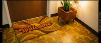 Retro welcome mat with colorful sunburst design on vintage tile floor near wooden door and mid-century modern plant stand with warm lighting.
