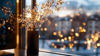 Dried flowers in ceramic vase on windowsill at dusk with blurred city lights creating magical bokeh effect through window glass. Cozy winter evening atmosphere.