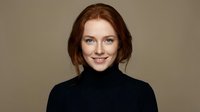 Professional redhead woman in black turtleneck sweater smiling confidently against neutral beige background for business and corporate marketing materials.