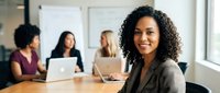 Confident mixed-race businesswoman smiling in modern office boardroom with diverse female colleagues working on laptops in background.