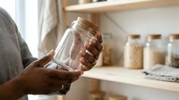 Empty glass jar held by person with dark skin tone in kitchen pantry with stored dry goods and wooden shelves for food storage organization.