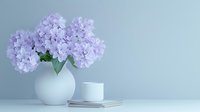 Delicate purple hydrangea flowers in white spherical vase against light gray wall, with minimalist white cup and stacked notebooks creating serene composition.