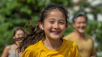 Young Hispanic girl with curly hair smiling joyfully in yellow shirt outdoors with blurred family in background during summer day activities.