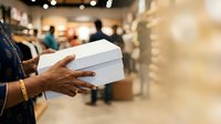 Woman holding white box in retail store with blurred shopping background, suitable for e-commerce, retail marketing, and customer service promotional materials.