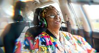 Happy elderly African American woman with glasses and colorful headwrap laughing joyfully while wearing headphones in bright interior setting.