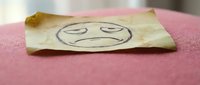 Crumpled yellow sticky note with hand-drawn sad face emoji on pink fabric surface expressing disappointment, frustration or negative emotions for office concepts.