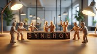 Wooden figurines standing around blocks spelling synergy on office desk with warm lighting and plants in background, representing teamwork and collaboration.