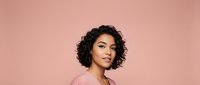 Young African American woman with curly hair wearing pink top against pink background, studio portrait for beauty and lifestyle content.