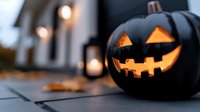 Black carved pumpkin jack-o-lantern with glowing orange face sits on wooden surface with warm lights and house in blurred background for Halloween decoration.