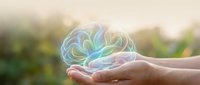 Glowing holographic brain floating above open palms in soft natural light representing mental health awareness, neuroscience research and cognitive wellness concepts.