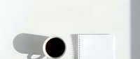Overhead view of steaming coffee mug casting shadow next to blank spiral notebook on white surface, minimalist workspace setup for design and lifestyle projects.
