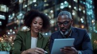 African American business colleagues reviewing digital tablet presentation outdoors at night, city lights and bokeh background create professional atmosphere.