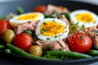 Fresh Nicoise-style salad with soft boiled eggs, tuna, green beans, cherry tomatoes and olives garnished with herbs and black pepper on dark plate.