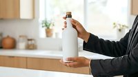 Person with dark skin tone holding white cleaning product bottle in modern kitchen environment for household cleaning purposes.