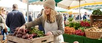 Young blonde woman in beige knit hat shopping for fresh vegetables at outdoor farmers market with colorful produce displays and shoppers in background.
