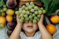 Green grapes cover the face of a young child with fair skin and brown hair, surrounded by fresh fruit including apricots and plums on white fabric.