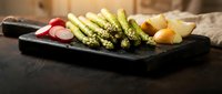 Grilled asparagus spears with radish slices and lemon wedges on dark slate serving board for healthy gourmet dining and restaurant presentation.