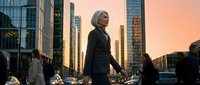 Mature woman with gray hair wearing black blazer walking through urban downtown street during golden hour sunset with modern skyscrapers and pedestrians.