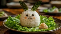 Cute bunny shaped rice ball with seaweed ears and carrot nose served on fresh lettuce leaves. Creative Asian food styling for children's meal or bento box lunch.
