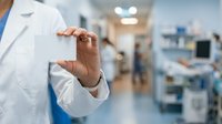 Doctor in white lab coat and stethoscope gesturing with hand in modern hospital corridor with blurred medical staff and equipment in background.
