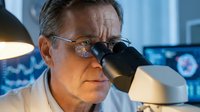 Senior Caucasian male scientist examining samples through microscope in modern laboratory wearing safety glasses and white coat for medical research.
