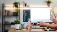 Hands holding blank white tablet device in modern home office workspace with shelving, plants, and natural lighting.
